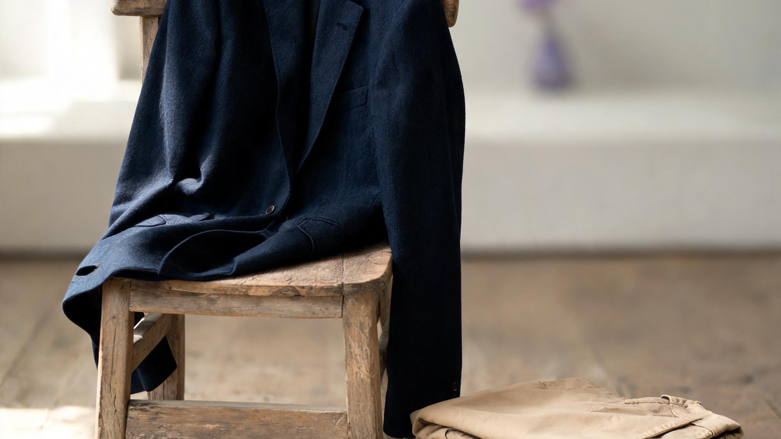 Alvin's Club — a navy blue unstructured blazer draped over a rustic wooden chair next to a pair of folded tan chinos